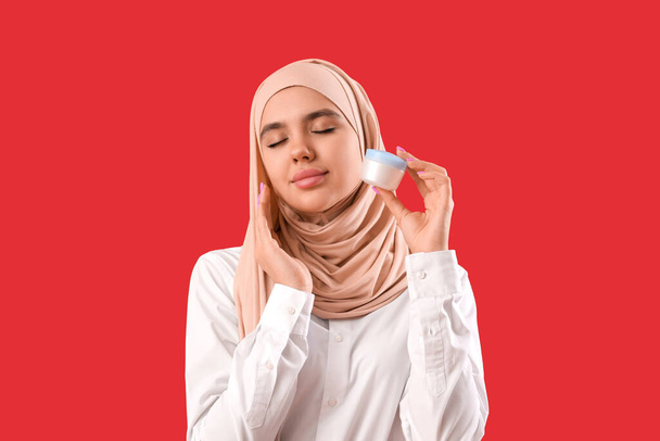 Kırmızı arka planda krema kavanozu olan tesettürlü genç Müslüman kadın. - Fotoğraf, Görsel
