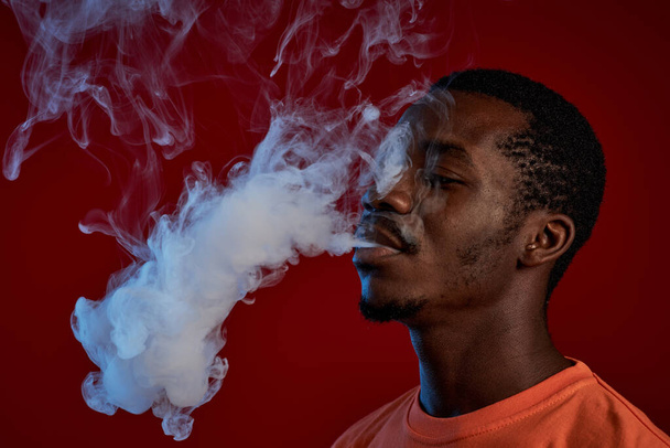 Jonge zwarte man in oranje t-shirt roken e-sigaret voor de camera tegen rode achtergrond en uitademen wolk van witte rook - Foto, afbeelding