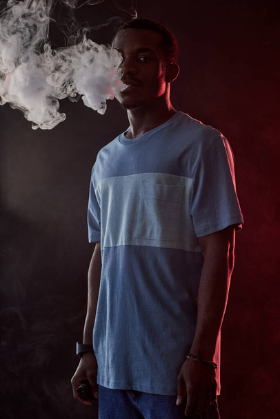 Młody Afroamerykanin w t-shircie i dżinsach z elektronicznym papierosem stojący przed kamerą i palący w ciemności - Zdjęcie, obraz