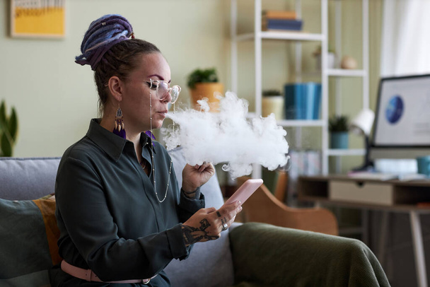 Jovem empresária rolando no celular e fumando cigarro eletrônico enquanto se senta no sofá no escritório em casa na pausa - Foto, Imagem