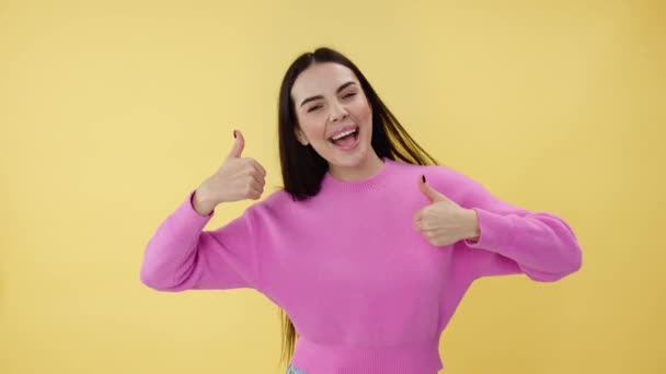Portret van een aantrekkelijke dame met bruin haar die vrolijk danst in de studio en duimen omhoog wijst. Blanke vrouw gekleed in roze slijtage uitstralen positiviteit en geven aanbeveling over gele achtergrond. - Video