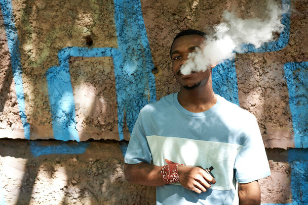Młody Afroamerykanin w koszulce stojący przy ścianie budynku przed kamerą i palący elektronicznego papierosa w letni dzień - Zdjęcie, obraz