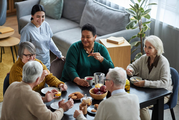 Высокий угол обзора группы пожилых людей, сидящих за столом вместе и счастливо улыбающихся в доме престарелых - Фото, изображение