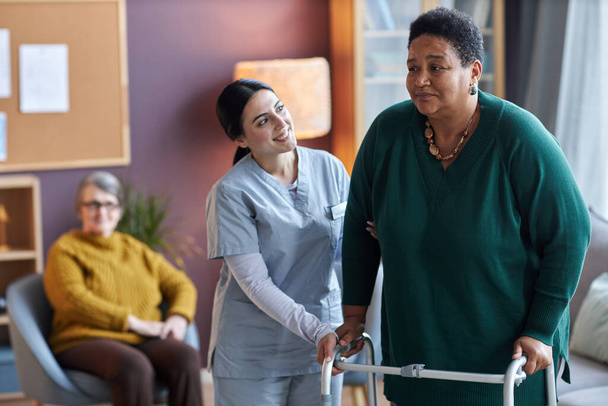 Portrait of smiling young nurse helping black senior woman using mobility walker in retirement home, copy space - Photo, Image