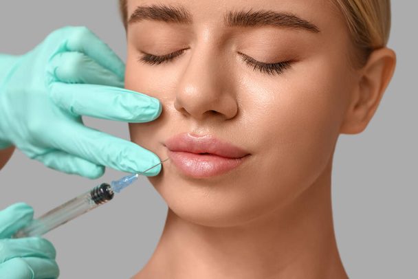 Mujer joven recibiendo inyección de relleno en labios sobre fondo gris - Foto, imagen