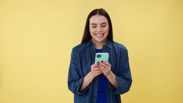 Αρκετά καυκάσια κυρία με τζιν πουκάμισο δακτυλογραφεί μηνύματα στο smartphone, ενώ στέκεται στο στούντιο. Χαρούμενη γυναίκα με μακριά καστανά μαλλιά έχοντας online επικοινωνία χρησιμοποιώντας σύγχρονο gadget πάνω από κίτρινο φόντο. - Πλάνα, βίντεο