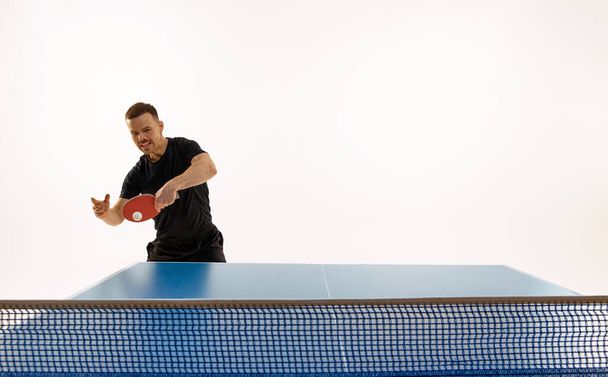 Siyah spor giyim giymiş atletik bir adam beyaz arka planda masa tenisi oynuyor. Agresif bir oyun tarzı. Profesyonel spor, dinamik ve enerji kavramı, şampiyonluk, turnuva. Ad. - Fotoğraf, Görsel
