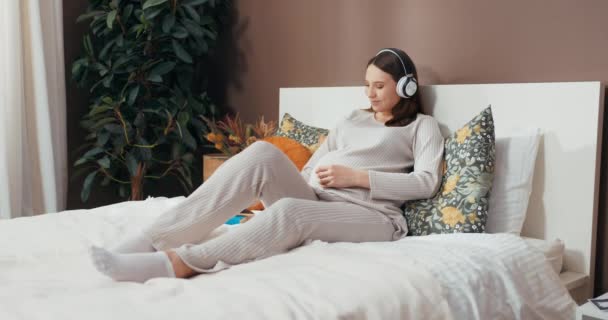 Ευτυχισμένο smiley όμορφη έγκυος γυναίκα βρίσκεται σε ένα κρεβάτι, κρατώντας την κοιλιά της και μιλώντας στο μωρό της, ενώ ακούγοντας ήρεμη μουσική μέσω ακουστικών κουβέντα με τα μέλη της οικογένειας . - Πλάνα, βίντεο