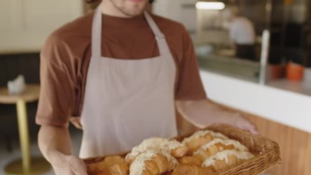 Retrato de inclinación hacia arriba tiro de joven hombre alegre en delantal sosteniendo bandeja de croissants recién horneados y posando para la cámara con sonrisa durante la jornada laboral en la panadería - Metraje, vídeo