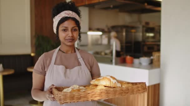 Porträt einer jungen glücklichen Frau in Schürze, die ein Tablett voller frischer Mandelcroissants in der Hand hält und in die Kamera lächelt, während sie in der Bäckerei arbeitet - Filmmaterial, Video