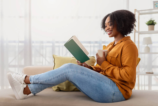 Eine entspannte afrikanisch-amerikanische Frau mit lockigem Haar und strahlendem Lächeln sitzt im Schneidersitz auf einer Couch, liest ein Buch und nippt an einer Tasse in einem gut beleuchteten, gemütlichen Wohnraum. - Foto, Bild