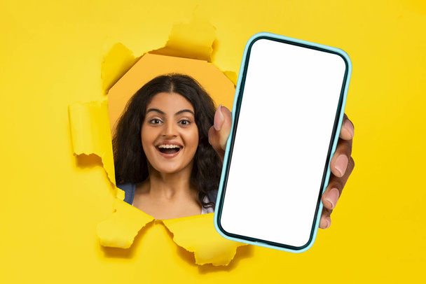 Feliz milenar senhora hindu segurando telefone na mão rompendo o pano de fundo de papel amarelo, apresentando tela em branco do celular, oferecendo maquete para promoção digital, recomendando aplicativo. Colagem - Foto, Imagem