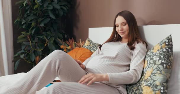Mujer embarazada hermosa sonriente de entre 20 y 30 años acostada en una cama, tocándose el vientre y escuchando música a través de auriculares con una expresión serena. - Metraje, vídeo