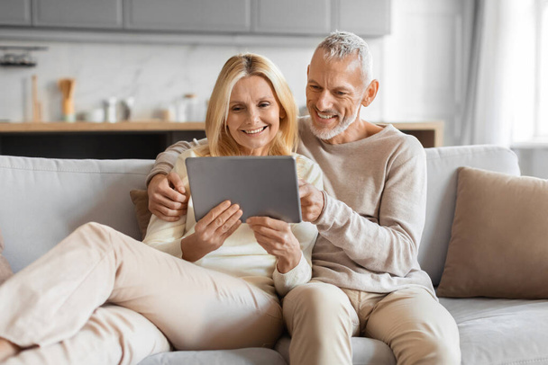 Pensioen en internet leuk concept. Opgewonden lachende oudere echtgenoten met behulp van digitale tablet computer, websurfen of film kijken online, rusten op de bank thuis, omarmen, vrije ruimte - Foto, afbeelding
