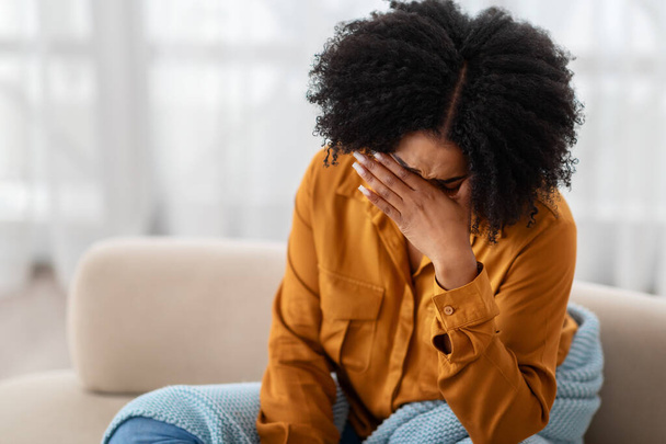 Una joven afroamericana triste con una camisa amarilla mostaza se sienta en un sofá beige, cubriéndose la cara con una mano, expresando una fuerte emoción de angustia, tristeza o frustración. - Foto, Imagen