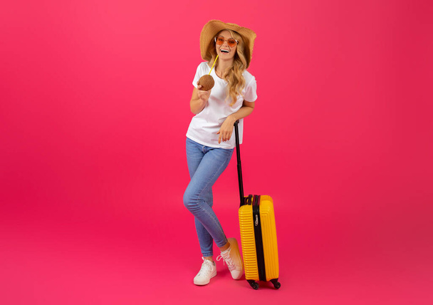 Dream Vacation. Joyful Young Blonde Woman Traveler With Yellow Suitcase Drinking Tropical Cocktail From Coconut, Posing Over Pink Studio Background, Full Length Shot Of Happy Tourist. Copy Space - Photo, Image