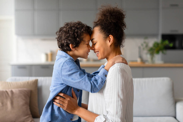 Preteen black curly-haired boy and his mother affectionately touching noses, sharing loving embrace in warmly lit home environment, sitting on couch, side view - Photo, Image