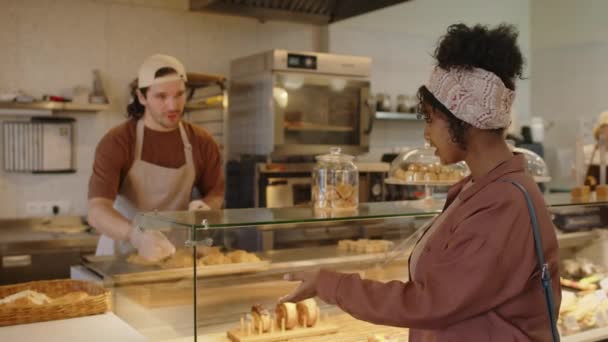 Jovem mulher escolhendo pastelaria e conversando com vendedor em avental e luvas ao visitar padaria - Filmagem, Vídeo