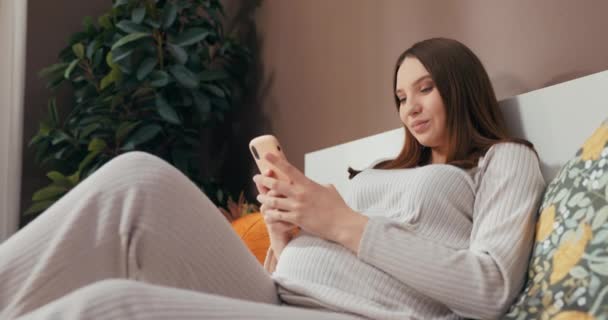 Ελκυστική νεαρή χαμογελαστή έγκυος γυναίκα βρίσκεται σε ένα κρεβάτι, κουβεντιάζοντας σε ένα κινητό τηλέφωνο με ένα μέλος της οικογένειας και χαμογελώντας χαρούμενα, ενώ χαϊδεύοντας το στομάχι της. - Πλάνα, βίντεο