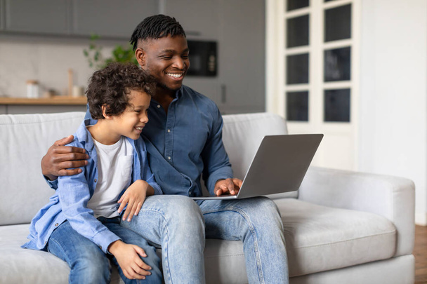 Hymyilevä musta isä ja iloinen esiteini-ikäinen poika istuvat mukavasti sohvalla, nauttivat aikaa yhdessä katsoessaan kannettavaa näyttöä viihtyisässä kotiympäristössä. - Valokuva, kuva