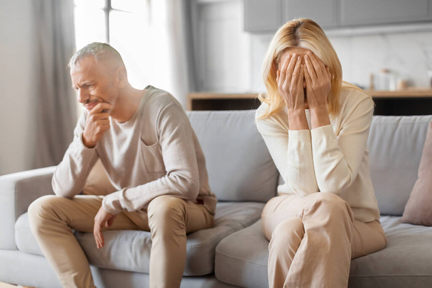 Mature blonde woman crying after quarrel with husband. Unhappy senior couple spouses husband and wife sitting on couch at home, thinking about divorce. Cheating, dishonesty, adultery in marriage - Photo, Image