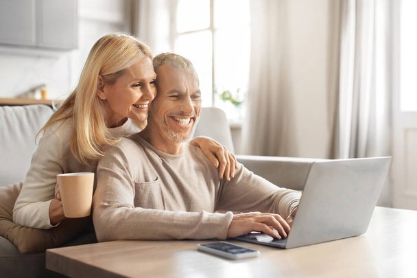 Pensioen en internet leuk concept. Opgewonden lachende oudere echtgenoten met behulp van moderne laptop computer, websurfen of films kijken online, rusten op de bank thuis, omhelzen, koffie drinken, vrije ruimte - Foto, afbeelding