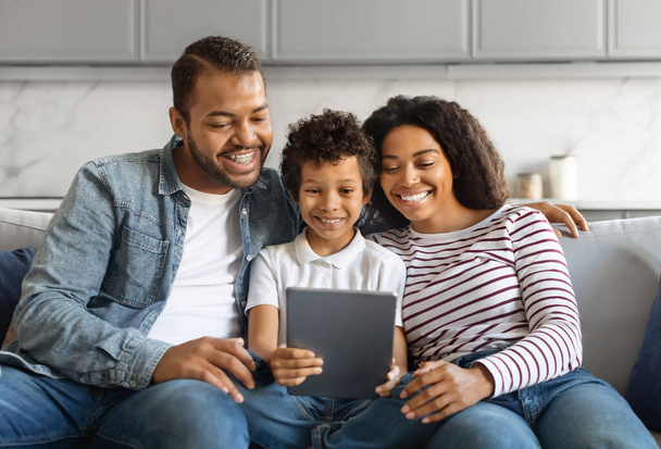 Famille noire avec fils préadolescent souriant joyeusement tout en utilisant la tablette numérique ensemble, heureux parents afro-américains et enfant mâle assis sur un canapé confortable à la maison, se détendre avec gadget moderne - Photo, image