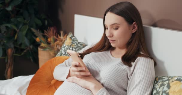 Ευτυχισμένη νεαρή χαμογελαστή έγκυος γυναίκα κρατώντας ένα κινητό τηλέφωνο πριν το αφήσει κάτω για να μιλήσει απαλά στο αγέννητο μωρό της, δημιουργώντας μια οικεία στιγμή αγάπης και σύνδεσης. - Πλάνα, βίντεο