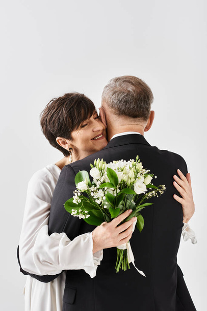 Een bruid en bruidegom van middelbare leeftijd in trouwjurken delen een oprechte knuffel om hun speciale dag in een studio te vieren. - Foto, afbeelding