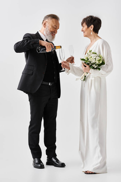Жених и невеста средних лет в свадебных платьях держат бокалы для шампанского, отмечают свой особенный день в студии. - Фото, изображение