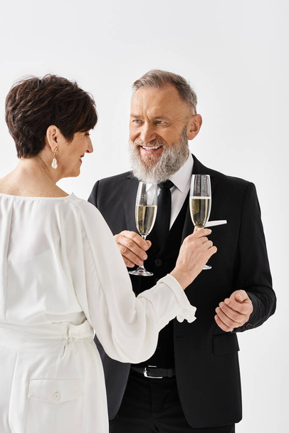 Novia y novio de mediana edad en traje de boda celebrando su día especial al criar flautas de champán en un entorno de estudio. - Foto, imagen