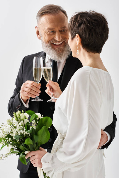 Novia y novio de mediana edad en traje de boda tintineo copas de champán en un entorno de estudio, celebrando su día especial. - Foto, imagen