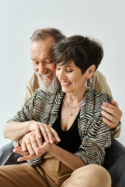 A middle-aged couple dressed stylishly, seated together on a chair, exuding grace and togetherness in a chic studio setting. - Фото, изображение
