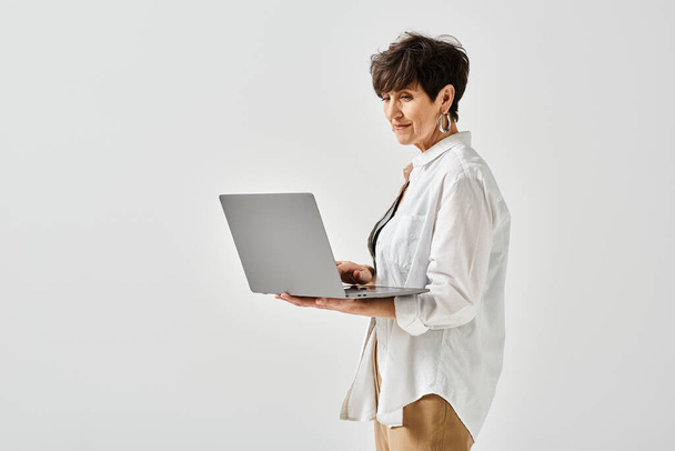 Elegante mujer de mediana edad con camisa blanca sosteniendo un portátil en un entorno de estudio. - Foto, Imagen