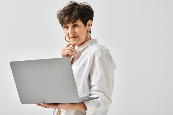 Kobieta w średnim wieku w stylowym stroju pewnie trzyma laptopa w rękach w studio.. - Zdjęcie, obraz