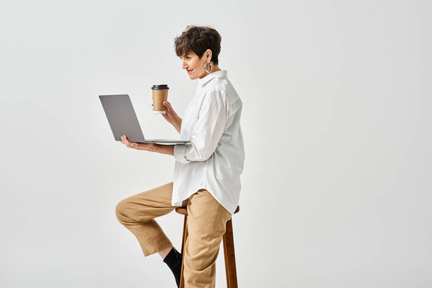 Une femme d'âge moyen en tenue élégante s'assoit sur un tabouret, travaillant sur un ordinateur portable dans un cadre de studio. - Photo, image
