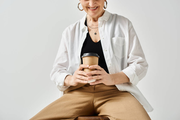 Een vrouw van middelbare leeftijd in stijlvolle kleding zit op een kruk, rustig een kopje koffie vasthoudend.. - Foto, afbeelding