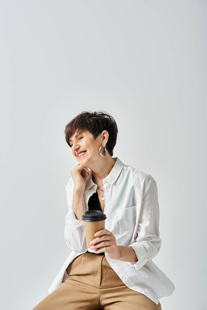 Una mujer de mediana edad, elegantemente vestida, con el pelo corto se sienta en un taburete, sosteniendo delicadamente una taza de café, perdida en el pensamiento. - Foto, imagen