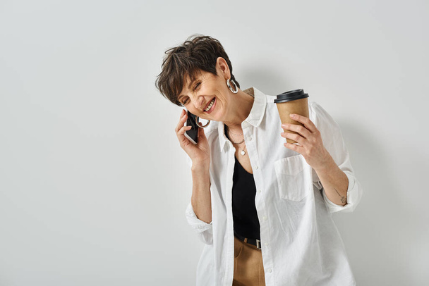 Una donna di mezza età con capelli corti multitasking, tenendo in mano una tazza di caffè e chattando sul cellulare in uno studio alla moda. - Foto, immagini