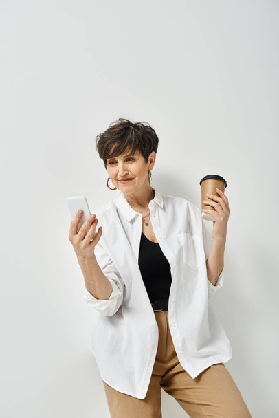 Stylowa kobieta w średnim wieku z krótkimi włosami trzyma filiżankę kawy w jednej ręce, a komórkę w drugiej.. - Zdjęcie, obraz