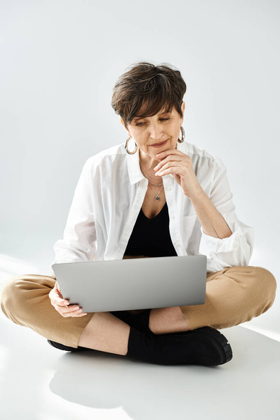 Una mujer de mediana edad elegantemente vestida con el pelo corto se sienta en el suelo, centrada en un ordenador portátil. - Foto, imagen