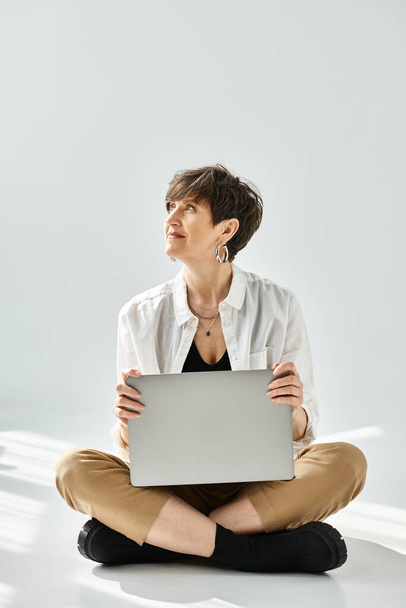 Kobieta w średnim wieku z krótkimi włosami ubrana stylowo, siedząca na podłodze i pracująca na laptopie w studio. - Zdjęcie, obraz