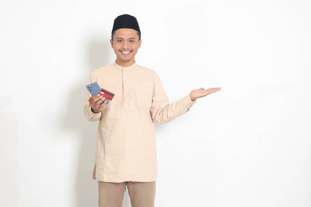 Retrato del atractivo hombre musulmán asiático en camisa koko con gorra de calavera sosteniendo un teléfono móvil y tarjeta de crédito mientras señala con el dedo hacia un lado. Imagen aislada sobre fondo blanco - Foto, imagen
