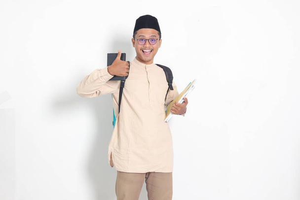 Portrait de l'étudiant asiatique musulman excité en chemise koko avec crâne portant sac à dos, tenant le livre de l'école, montrant pouce vers le haut geste. Concept d'éducation islamique. Image isolée sur fond blanc - Photo, image