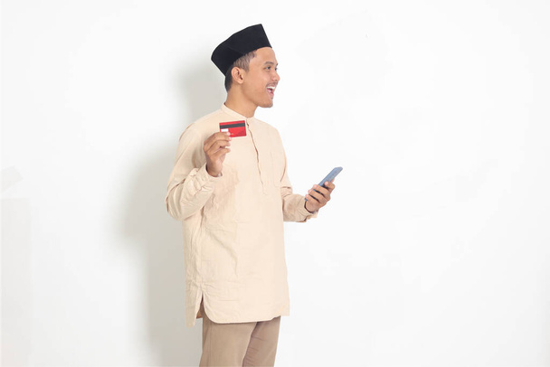 Porträt eines attraktiven asiatischen muslimischen Mannes im Kokohemd mit Totenkopf, der ein Mobiltelefon in der Hand hält und seine Kreditkarte präsentiert. Isoliertes Bild auf weißem Hintergrund - Foto, Bild