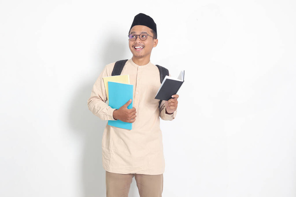 Portrait of excited student Asian muslim man in koko shirt with skullcap carrying school books, while reading his notebook. Isolated image on white background - Photo, Image
