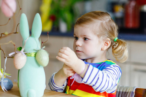 Cute little little maluch dziewczyna dekorowanie drzewa i króliczek z kolorowych pastelowych jaj plastikowych. Szczęśliwe dziecko bawiące się dekoracjami wielkanocnymi. Uroczy zdrowy uśmiechnięty dzieciak w korzystaniu z rodzinnych wakacji. - Zdjęcie, obraz