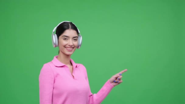 Zadowolona młoda, kaukaska kobieta nosząca słuchawki skierowane prosto na palec, uśmiechnięta i patrząca w kamerę, stojąca nad zielonym tłem. Przestrzeń kopiowania. - Materiał filmowy, wideo