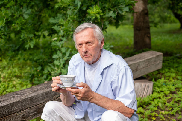 Starší muž sedí sám na dřevěné lavičce v zahradě a drží šálek kávy. Klidné prostředí vyjadřuje mírumilovnou, kontemplativní náladu, vhodnou pro wellness témata. Kvalitní fotografie - Fotografie, Obrázek