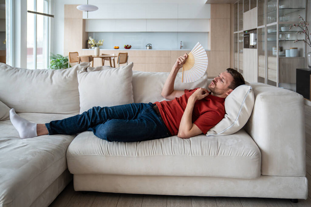 Ylikuumentunut hikoileva mies makaa sohvalla, vilkuttaa käsi tuuletin, kärsivät korkeassa lämpötilassa huoneessa, koska rikki hoitoaine, puute tuuletin, ilmanpuhallin. Miesten huonovointisuus, kuivuminen - Valokuva, kuva